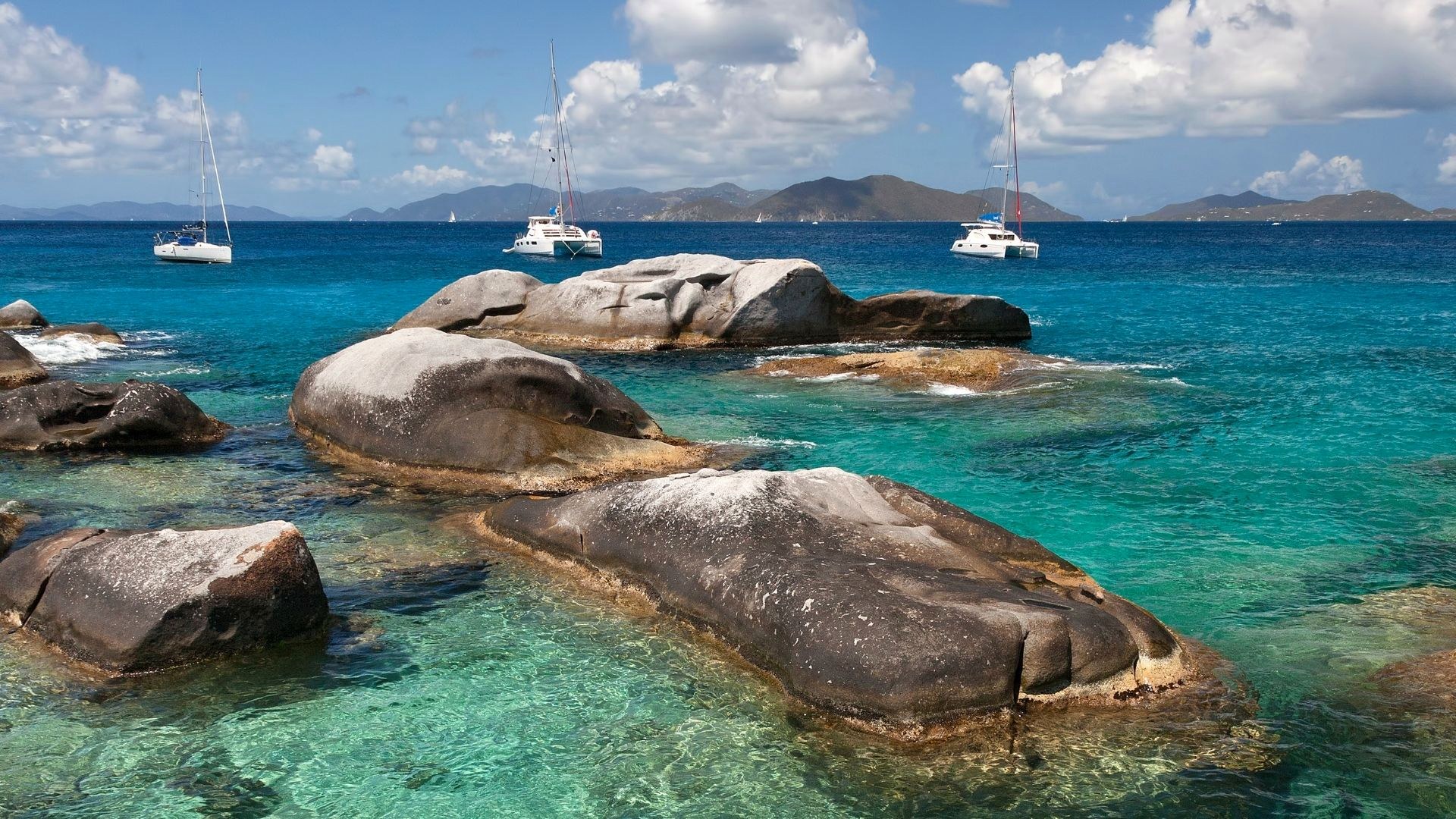 Runde Granitfelsen in glasklarem türkisem Wasser, dahinter Katamarane vor Anker und die Inseln der British Virgin Islands